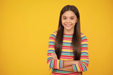 Happy face, positive and smiling emotions of teenager girl. Copy space. Cute little teenager girl crossing arms over yellow background