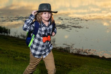 Summer holiday. Child with binoculars travelling outdoors. Boy traveler with backpack in a summer day. Portrait of a little boy exploring wildlife. Hiking and adventure concept