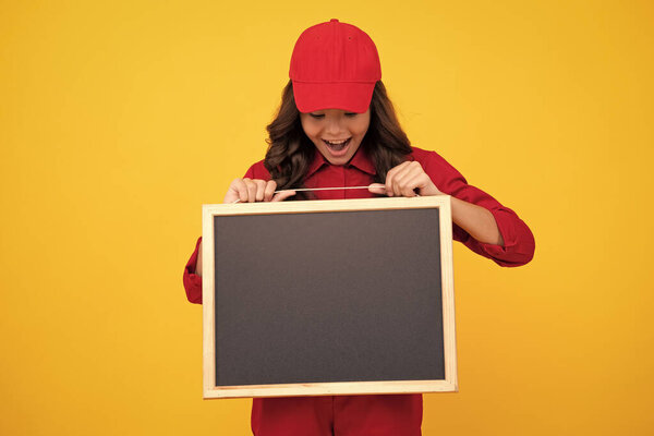 Excited child girl in red uniform and cap hold blackboard. Teenage girl hold blackboard isolated on yellow background. Copy space on empty board, mock up. Excited teenager girl