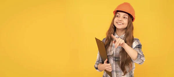 stock image Builder teenager girl in helmet. happy teen girl in helmet and checkered shirt making notes on clipboard, busy. Child in hard hat horizontal poster design. Banner header, copy space