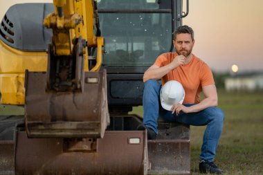 Kafkasyalı bir adam, inşaat alanında miğferli bir inşaat işçisi. Buldozer ya da kazıcı traktörünün yanındaki sanayi mühendisi. İnşaat endüstrisi kavramı. İnşaat alanı yöneticisi