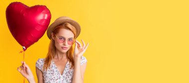 happy girl in sunglasses hold love heart balloon on yellow background. Woman isolated face portrait, banner with copy space