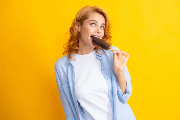 Young woman eat ice creams with chocolate glaze on yellow background. Funny redhead woman with ice cream