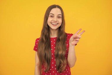 Cheaky teen girl giving victory sign. Happy teenager child smiling with v-sign, positive gesture, standing against yellow isolated background