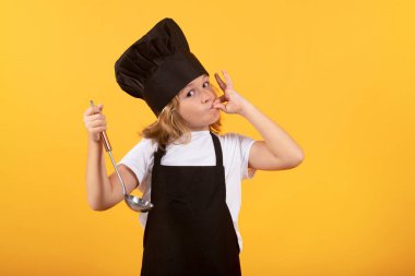 Mutfak kepçesiyle aşçı çocuk, stüdyo portresi. Çocuk pişirme, küçük şef yemek hazırlıyor. Sarı stüdyodaki aşçı şapkalı ve önlüklü çocuk izole edilmiş.