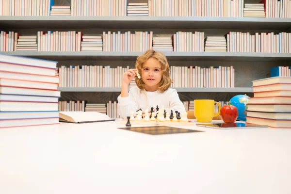 Chess school. Concentrated kid play chess. Smart child play chess in the library by the bookshelves. Educational concept. Boy thinking about chess. The concept of learning and growing children