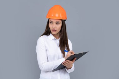 Serious woman worker in protective helmet with clipboard isolated on grey background. Young woman construction manager. Architect woman, female worker in hardhat helmet