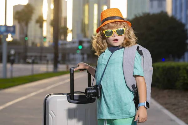 Child traveler standing and holding suitcase outdoor. Tourist kid boy having cheerful holiday trip. Child travel with travel bag. Child with suitcase dreams of travel, adventure, vacation