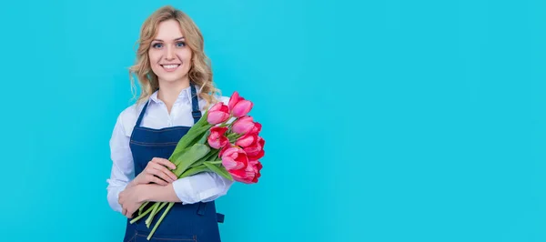 positive flower seller woman in apron with spring tulip flowers on blue background. Woman isolated face portrait, banner with mock up copy space