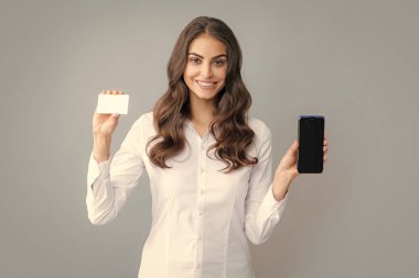 Amazed young brunette woman in shirt using mobile cell smart phone hold credit bank card isolated on gray background studio portrait