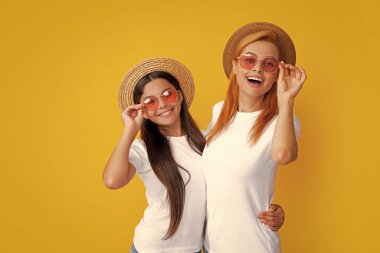 Happy stylish mother and daughter posing at studio yellow background, wearing straw hat and sunglasses. Summer family portrait