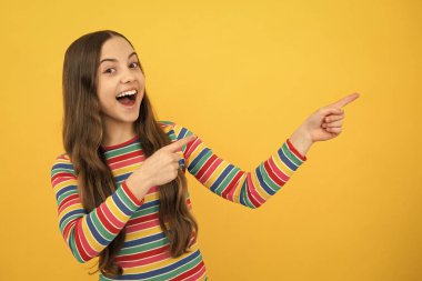 Close up portrait of teenager child girl showing at copy space, pointing to ads advertising, isolated over yellow background. Mock up copy space. Excited face, cheerful emotions of teenager girl