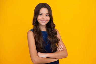 Mutlu kız yüzü, pozitif ve gülümseyen duygular. Kollarını kavuşturmuş genç bir kız, kameraya bakıyor, sarı stüdyo arka planında izole edilmiş.