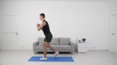 Fitness sporcusu ters hamleler yapıyor. Formda sporcu dengelemek için ön bacağına ağırlık veriyor. Evde spor egzersizleri var. Evde spor yapma rutini. Evde fitness var. Spor ve zindelik.