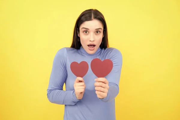 Girl feeling love. Portrait of attractive lovely cheerful girl holding in hands paper heart, isolated over yellow background. Love valentine concept