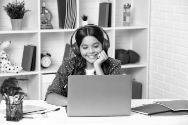 A young school girl student with wireless headphones sitting at the table, using laptop when studying. Distance learning, homeschooling, remote education.