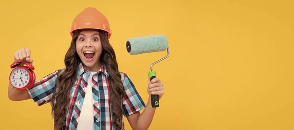 stock image amazed teen girl with curly hair in helmet hold paint roller and alarm clock, time to upgrade. Child builder in helmet horizontal poster design. Banner header, copy space