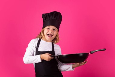 Çocuk aşçı tavayla yemek yapıyor. Aşçı çocuk, izole stüdyo geçmişiyle yemek hazırlıyor. Çocuklar yemek yapıyor. Önlüğü ve aşçı şapkası olan genç çocuk sağlıklı bir yemek hazırlıyor.