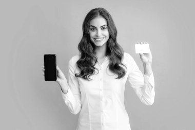 Amazed young brunette woman in shirt using mobile cell smart phone hold credit bank card isolated on gray background studio portrait