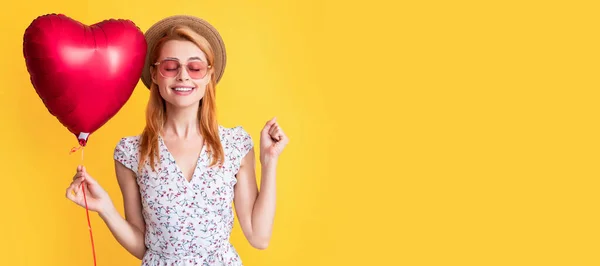 stock image glad young woman in straw hat and sunglasses hold love heart balloon on yellow background. Woman isolated face portrait, banner with copy space