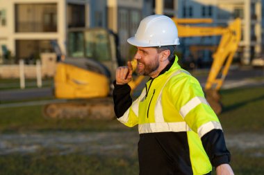 Caucasian man, construction worker in helmet at construction site. Industry engineer worker in hardhat near bulldozer or digger tractor. Concept of construction industry. Construction site manager