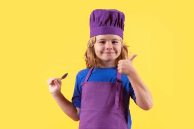 Komik çocuk aşçı kaşıkla aşçı, stüdyo portresi. Aşçı bozuntusu aşçı. Önlüklü ve aşçı şapkalı çocuk stüdyo izole edilmiş arka planda sağlıklı bir yemek hazırlıyor. Yemek pişirme süreci