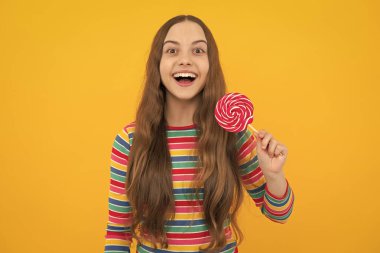 Funny child with lollipop over yellow isolated background. Sweet childhood life. Teen girl with yummy caramel lollipop, candy shop. Teenager with sweet sucker