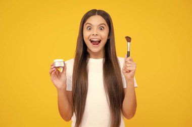 Güzellik, makyaj ve çocukların makyaj malzemeleri. Yüzüne kızaran fırçalı ergen bir kız çocuğu arka planda izole edilmiş. Heyecanlı bir genç, şaşkın ve neşeli duygular içinde