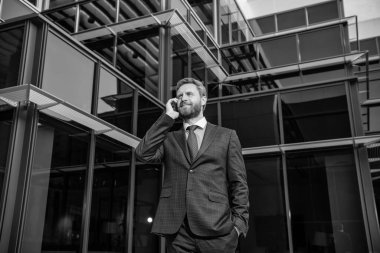 Gülümseyen, resmi giyinmiş, açık havada konuşan, modern iletişim kuran olgun bir iş adamı..