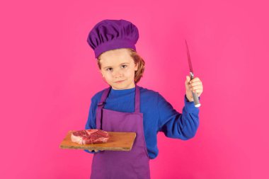 Çocuk aşçı et bifteği ve bıçakla kesme tahtasını tutuyor. Yemek pişiren çocuklar. Aşçı bozuntusu aşçı. Önlüklü ve aşçı şapkalı çocuk mutfakta sağlıklı bir yemek hazırlıyor. Yemek pişirme süreci