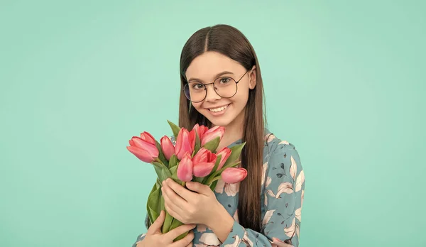 kid hold flowers for 8 of march. teen girl with spring bouquet on blue background. floral present. cheerful child in eyeglasses with tulips. mothers or womens day.
