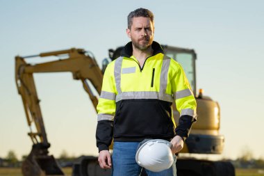 Man builder near excavator. Builder worker with excavator. Builder in helmet. Worker in hardhat. Portrait mechanical worker in construction helmet. Engineer builder foreman or repairman
