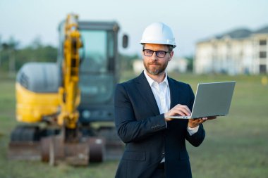 İnşaat alanında kask ve takım elbiseli bir inşaat yöneticisi. İnşaat müdürü ya da yönetici evin önünde şapka takıyor. Kazıcının yanındaki inşaat müdürü. Yenileme