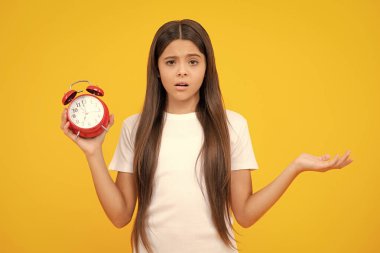 Çocuk saati sarı stüdyo arka planında izole edilmiş. Çalar saati olan genç bir çocuk saati gösteriyor. Kızgın yüz, üzgün genç kız duyguları.