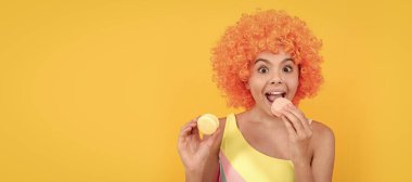 Mayo giyen, turuncu kıvırcık saçlı, sarı arka planda Fransız makaronu yiyen, diş bakımı yapan mutlu çocuk..