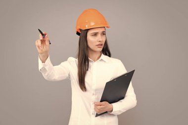 Miğferli mimar kadın işaret ediyor. Kasklı genç kadın gri arka planda pano tutuyor.