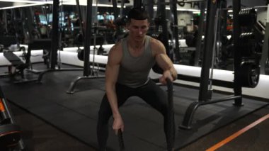 Spor salonunda savaş halatıyla Crossfit antrenmanı yapan aktif bir sporcu. Spor salonunda çapraz formda savaş ipi olan bir sporcu. Spor salonunda çapraz formundaki savaş ipi ekipmanlarıyla sporcu.