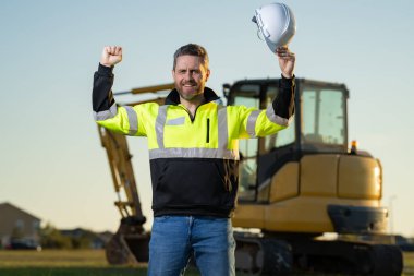 Man builder near excavator. Builder worker with excavator. Builder in helmet. Worker in hardhat. Portrait mechanical worker in construction helmet. Engineer builder foreman or repairman