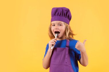 Komik çocuk aşçı kaşıkla aşçı, stüdyo portresi. Çocuk aşçı, izole stüdyo geçmişinde yemek hazırlıyor. Çocuklar yemek yapıyor. Önlüğü ve aşçı şapkası olan genç çocuk sağlıklı bir yemek hazırlıyor.