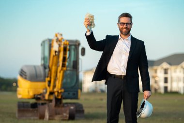İnşaat alanında mimar. Modern ev inşaatında kask ve takım elbiseli mimar. Güvenlik yeleği ve takım elbisesi olan bir mimar. Evin arka planında duran kendine güvenen mimar.