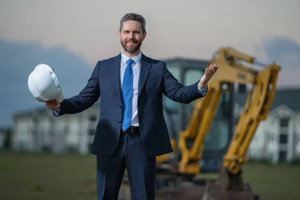 Construction business owner. Man in suit and hardhat halmet at building construction site. Civil engineer worker in front of house near excavator. Architect, supervisor, investor, construction manager