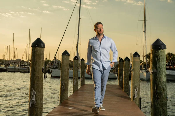 Yatta kendine güvenen yakışıklı bir adamın portresi. Yat kulübü. Moda gömlekli seksi adam yatın yanında poz veriyor. Şehirde başarılı bir erkek manken. Beyaz, orta yaşlı bir adam lüks yatın yanında.