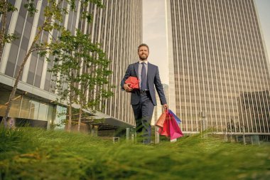 Formalite içinde başarılı bir adam alışveriş çantaları ve hediye kutuları ile ofis dışında yürüyüş yapar..