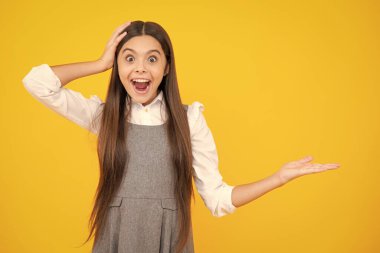 Close up portrait of teenager child girl showing at copy space, pointing to ads advertising, isolated over yellow background. Mock up copy space. Excited face. Amazed expression, cheerful and glad