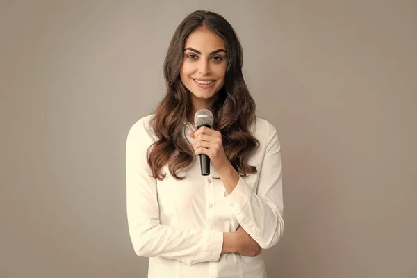 Beautiful business woman is speaking on conference. Stylish girl singing songs with microphone, holding mic at karaoke, posing against gray background