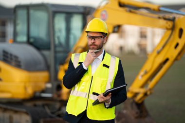 İnşaat alanında kask ve takım elbiseli bir inşaat yöneticisi. İnşaat müdürü ya da yönetici evin önünde şapka takıyor. Kazıcının yanındaki inşaat müdürü. Yenileme