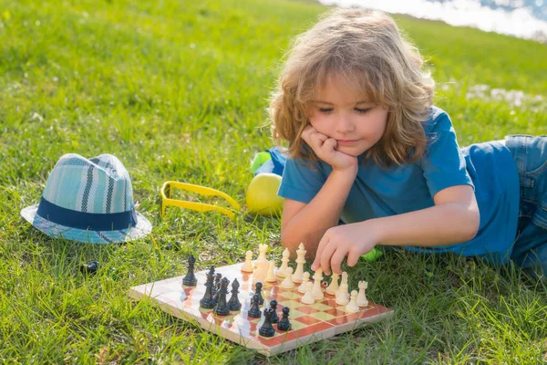 Concentrated child boy developing chess strategy, playing board game in backyard, laying on grass