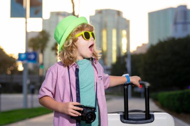 Seyahat çantası taşıyan bir çocuk turist. Elinde bavul olan bir çocuk şehir caddesinde yürüyor. Günlük giysiler ve şapka taşıyan gezgin turist çocuk.