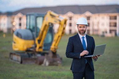 İnşaat alanında mimar. Modern ev inşaatında kask ve takım elbiseli mimar. Güvenlik yeleği ve takım elbisesi olan bir mimar. Evin arka planında duran kendine güvenen mimar.