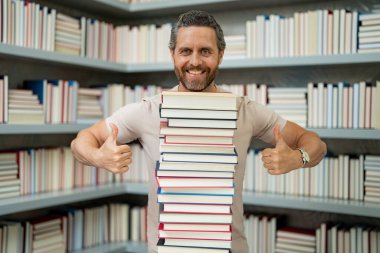 Kütüphane sınıfındaki öğretmenin portresi. Üniversite kütüphanesinde öğretmenler. Öğretmenler Günü. Komik öğretmen bir sürü kitap tutar. Kitapları olan çılgın bir öğretmen. Okul kitaplığındaki heyecanlı öğretmen